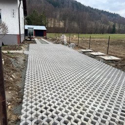 PolBel - Plus Dawid Grzanka - Ułożona szara ażurowa kostka brukowa tworząca podjazd na posesji. W tle dom, budynek gospodarczy, teren przygotowany pod ogrodzenie panelowe i zbocze góry.