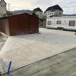Ukończony podjazd z jasnoszarej kostki brukowej, obok garaż blaszany i przyczepa kempingowa. Widoczna ubijarka. Brzeg podjazdu jeszcze nie zasypany ziemią.