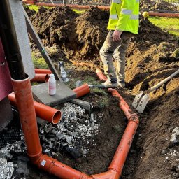PolBel - Plus Dawid Grzanka - Montaż rur kanalizacyjnych w wykopie w Laskach. Widoczne pomarańczowe rury, ziemia, narzędzia i pracownik w odblaskowej kamizelce. Prace ziemne w toku.