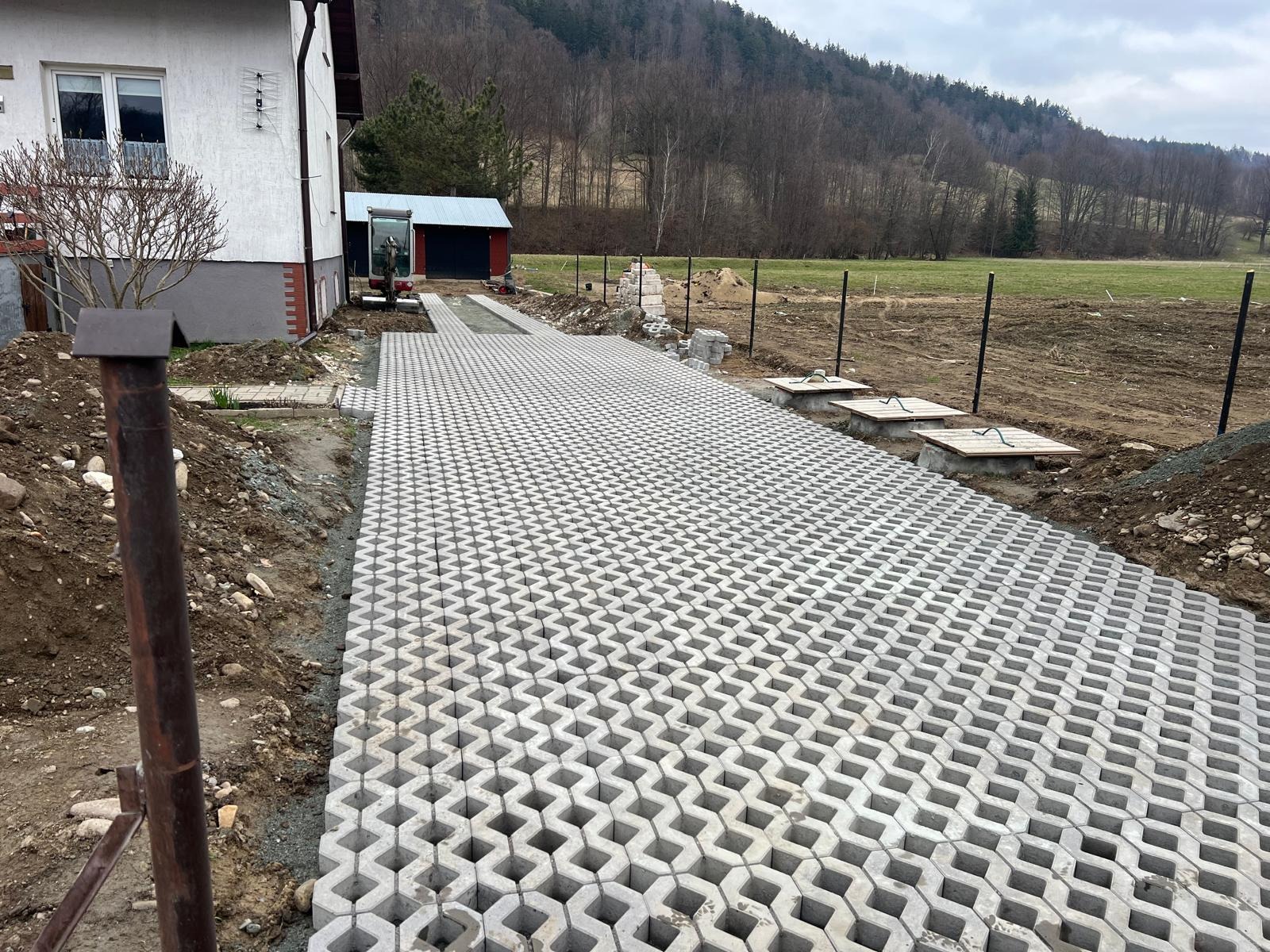 Ułożona szara ażurowa kostka brukowa tworząca podjazd na posesji. W tle dom, budynek gospodarczy, teren przygotowany pod ogrodzenie panelowe i zbocze góry.