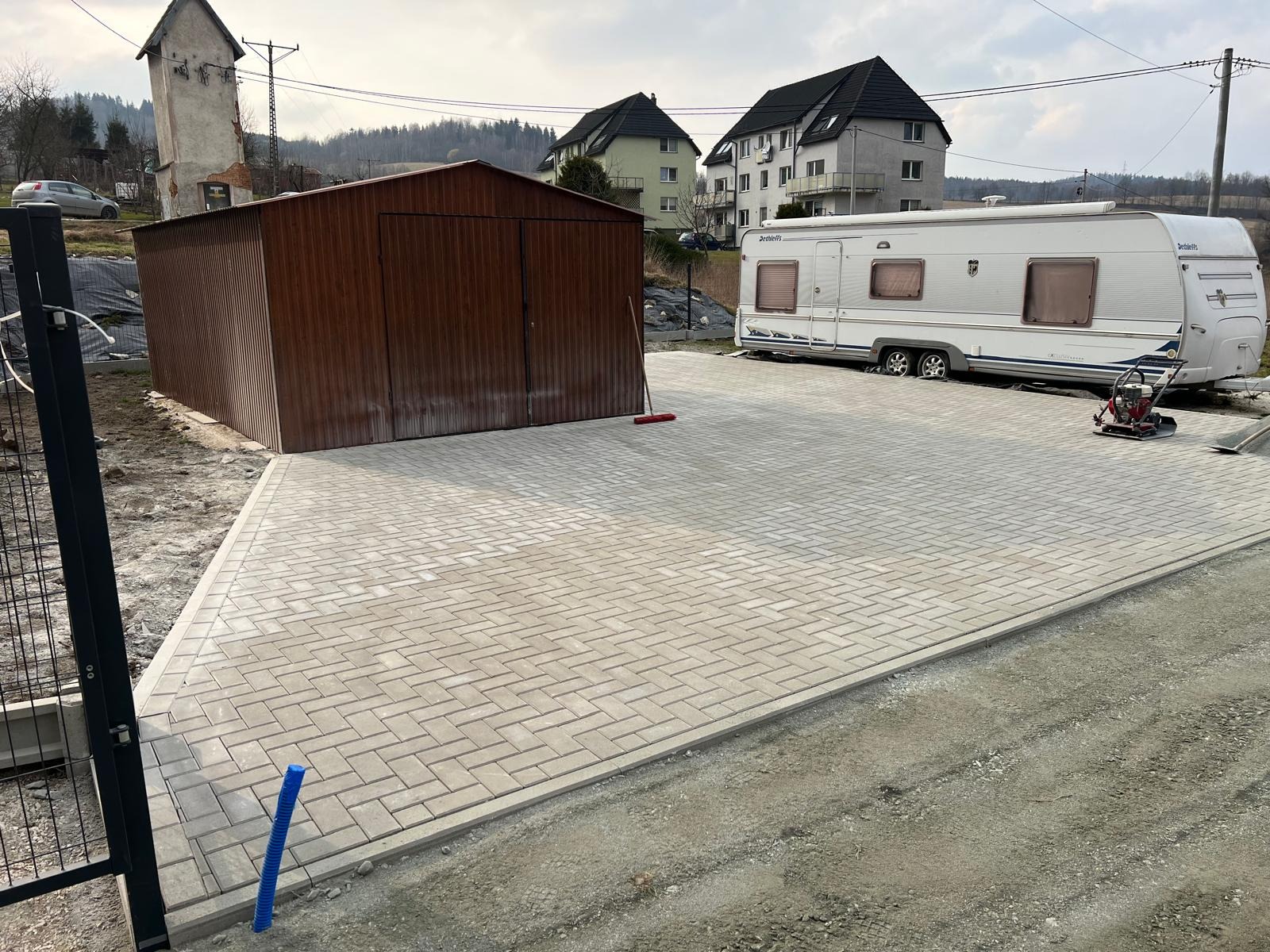 Ukończony podjazd z jasnoszarej kostki brukowej, obok garaż blaszany i przyczepa kempingowa. Widoczna ubijarka. Brzeg podjazdu jeszcze nie zasypany ziemią.