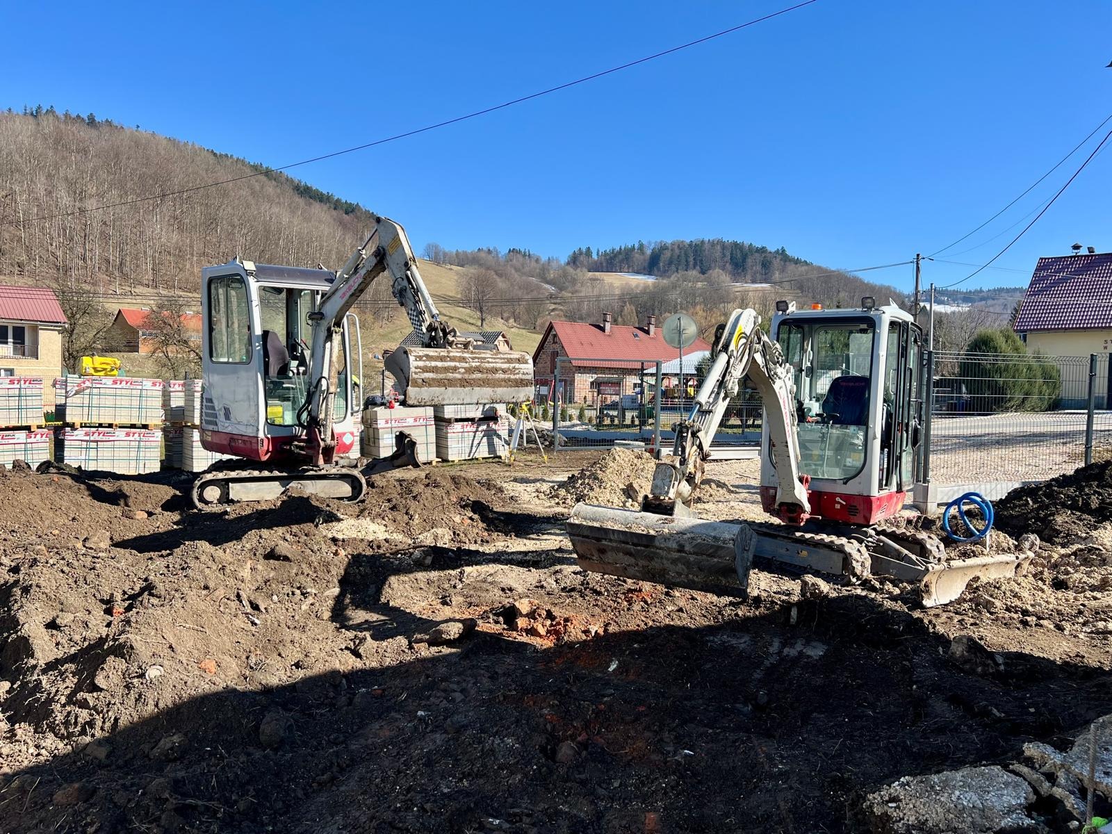 Dwie małe koparki na placu budowy, w tle ułożona kostka brukowa i budynki w oddali. Słoneczny dzień, błękitne niebo. Prace ziemne w toku.