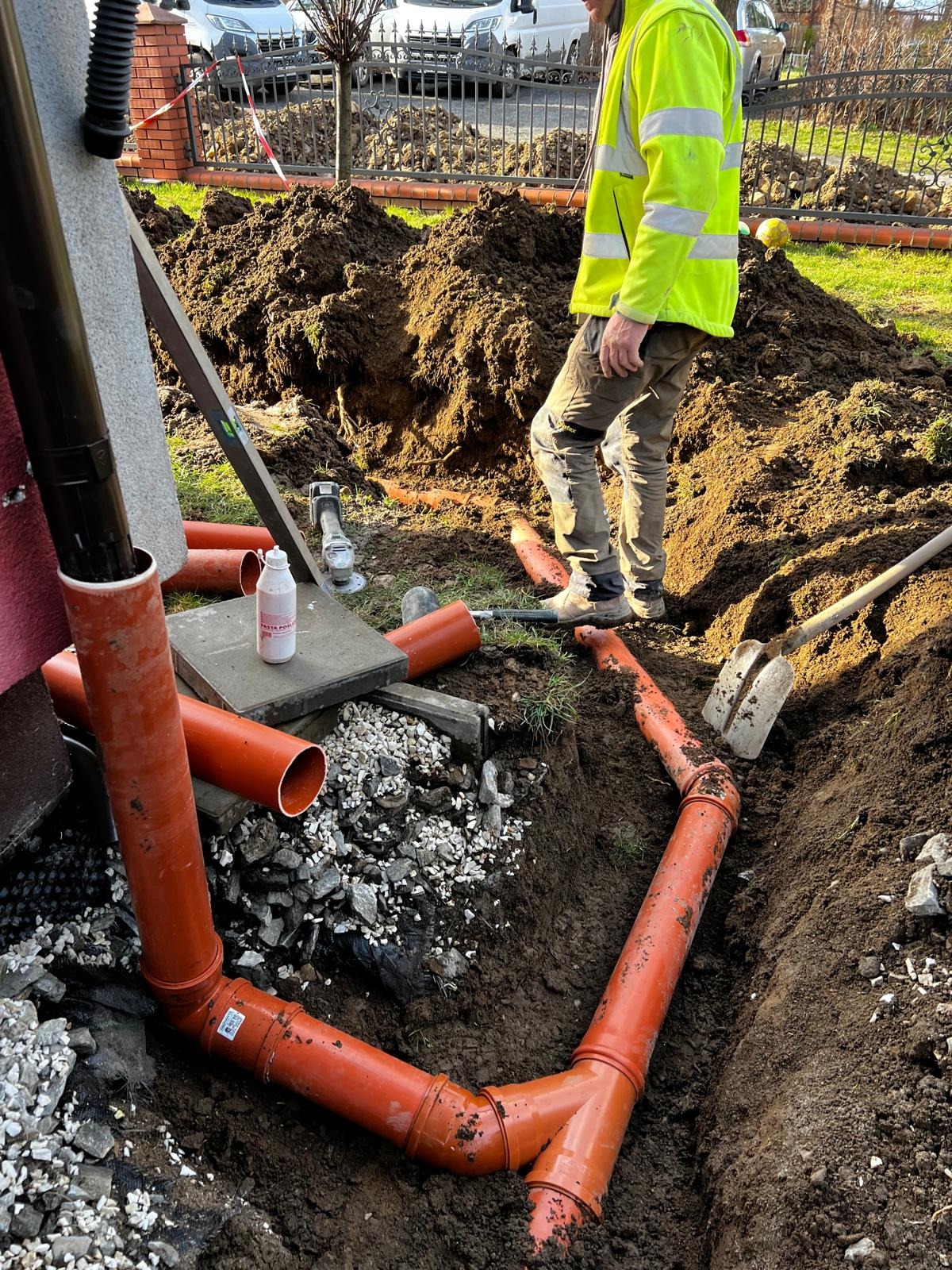 Montaż rur kanalizacyjnych w wykopie w Laskach. Widoczne pomarańczowe rury, ziemia, narzędzia i pracownik w odblaskowej kamizelce. Prace ziemne w toku.