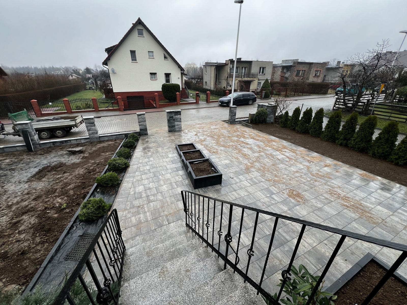 Ułożona kostka brukowa w odcieniach szarości na podjeździe, z elementami zieleni i czarnymi donicami. Widok z góry ze schodów, w tle domy i pochmurne niebo.