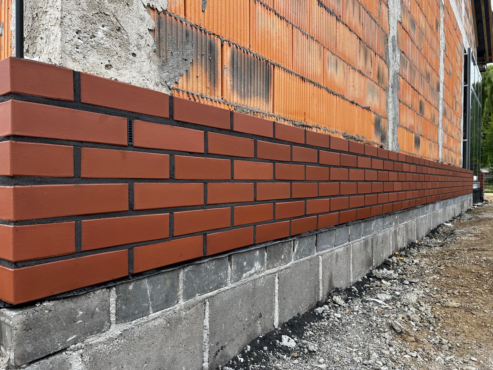 Ceglana elewacja klinkierowa z czarną fugą, wykończona na fundamencie budynku. Widoczne pomarańczowe bloczki na ścianie powyżej i gruz budowlany u podstawy.