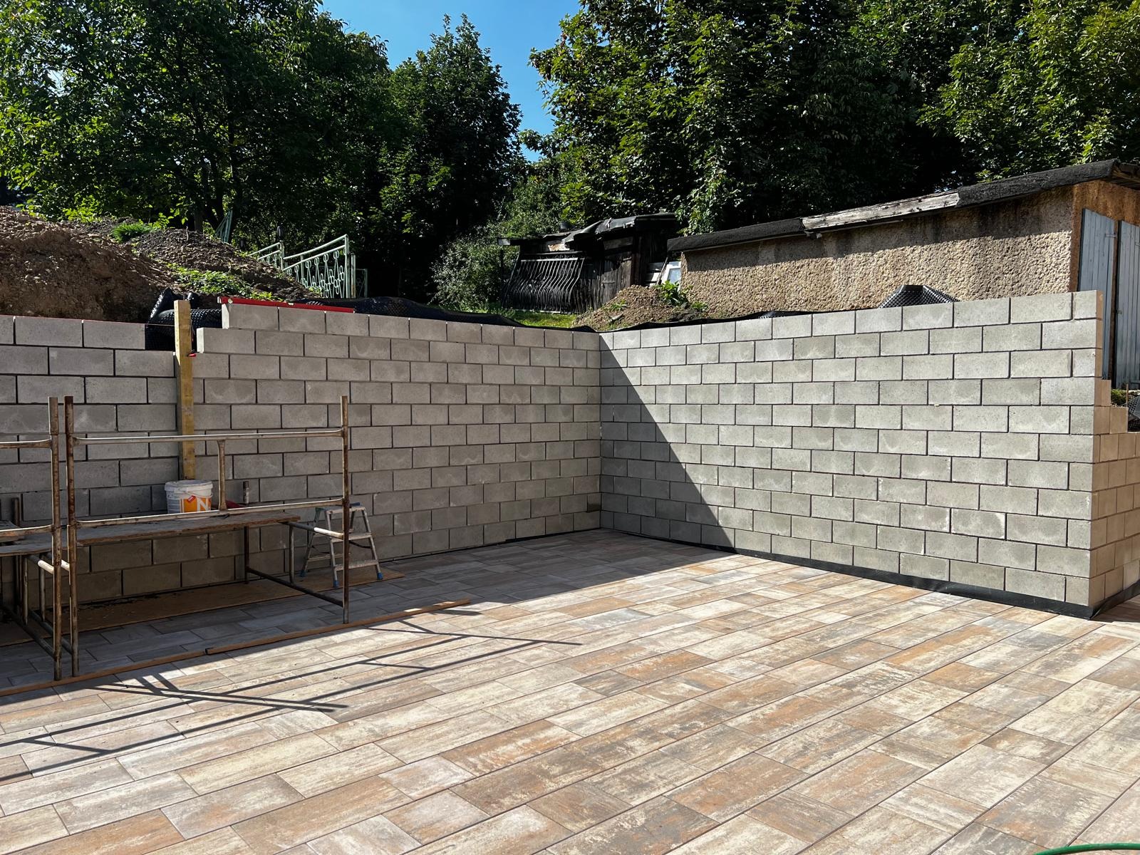 Wznoszenie muru oporowego z bloczków betonowych na posesji. Ułożona kostka brukowa. Widoczny rusztowanie i narzędzia. Tło: zieleń drzew.