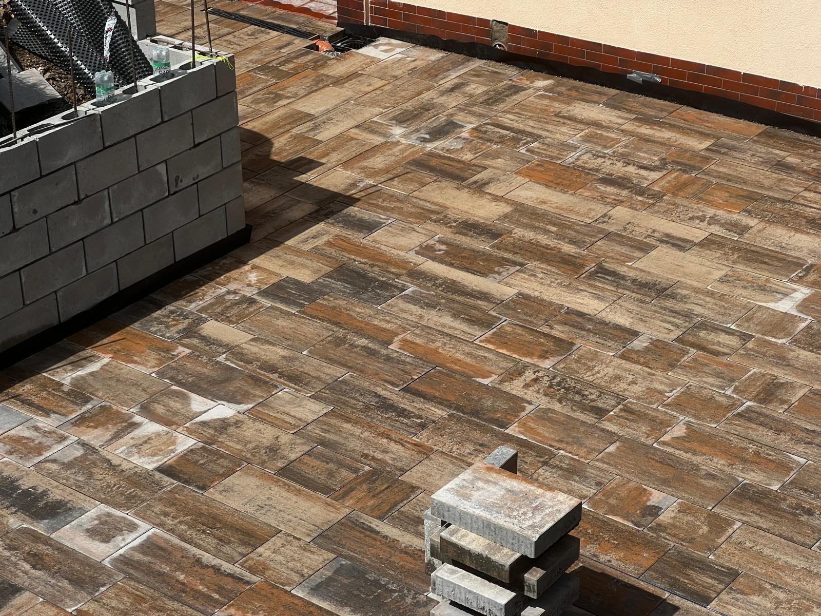 Ułożona kostka brukowa imitująca drewno, z fragmentem muru i stosem kostek. Widok z góry, ciepłe oświetlenie.