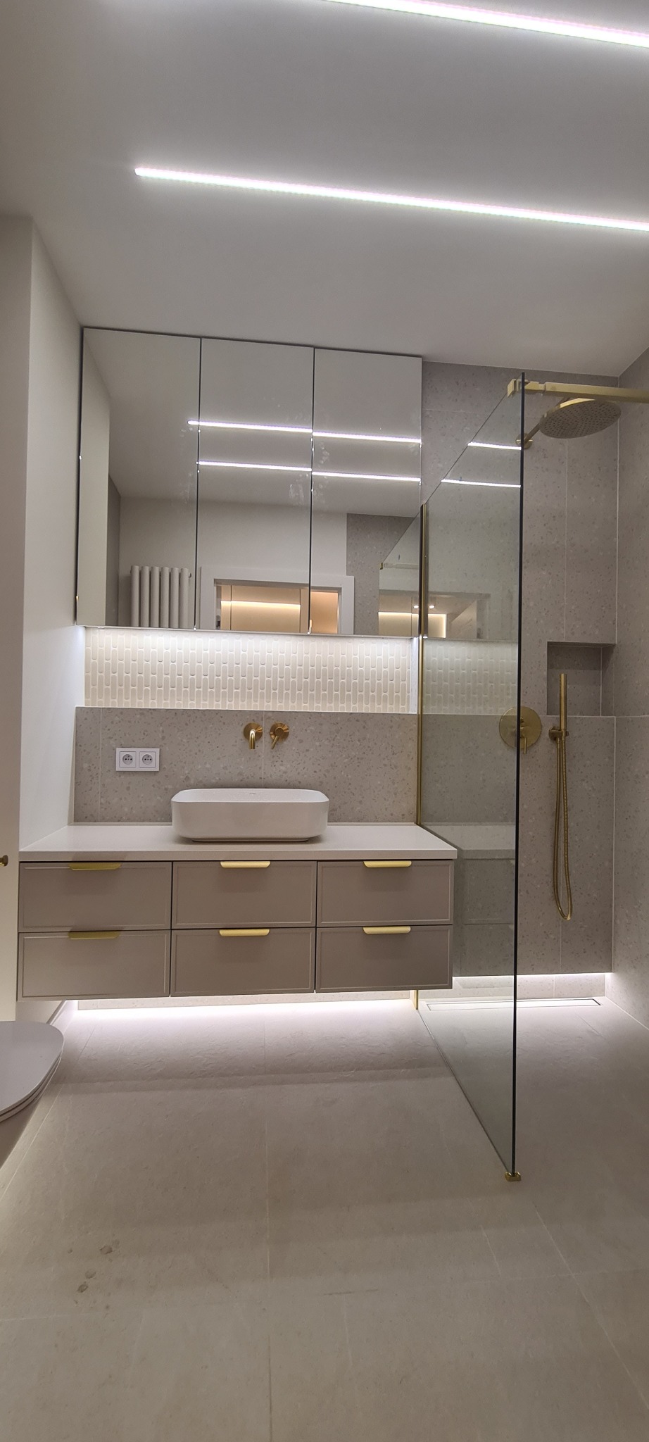 Elegancka łazienka z beżową szafką pod umywalką, złotą armaturą i kabiną prysznicową ze szkła. Oświetlenie LED podkreśla nowoczesny design.