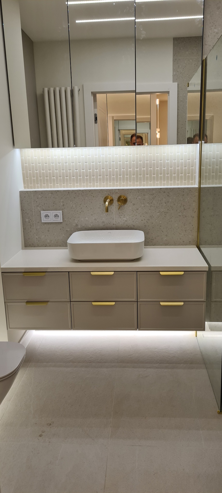 Jasna łazienka po remoncie: biała umywalka nablatowa, złota armatura, mozaika nad blatem, podświetlane szafki, lustra w zabudowie. Nowoczesne wykończenie wnętrza.