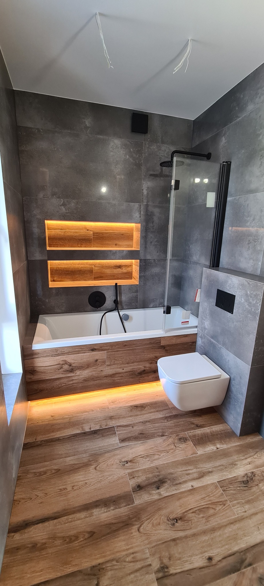 Nowoczesna łazienka z drewnopodobną podłogą, szarą glazurą, podświetlaną wanną i półkami wnękowymi. Uwagę zwraca minimalistyczna toaleta i czarna armatura.