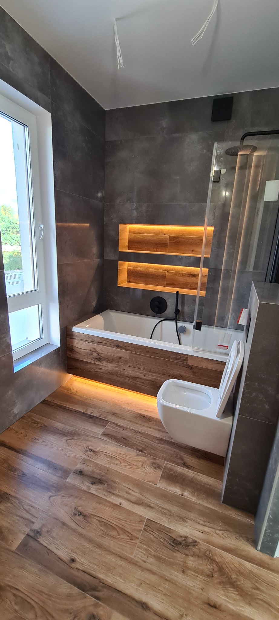 Nowoczesna łazienka z wanną obudowaną drewnem i podświetleniem LED, szare płytki, drewnopodobna podłoga, wisząca toaleta i kabina prysznicowa.