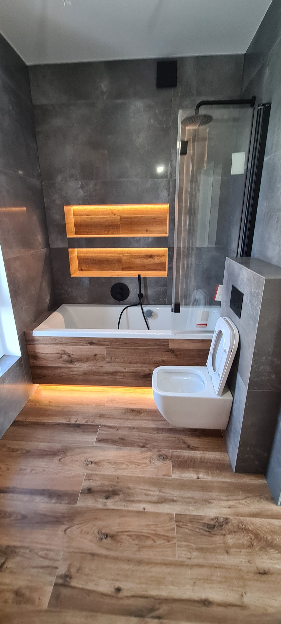 Nowoczesna łazienka w szarościach z drewnianymi akcentami. Podświetlana wanna i półki w ścianie, wisząca toaleta. Drewnopodobna podłoga, minimalistyczny design.