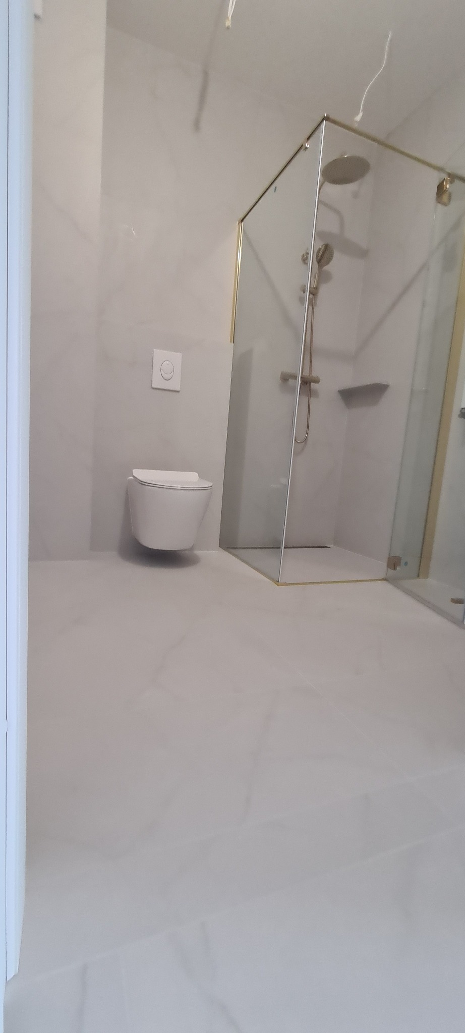 Nowoczesna łazienka z kabiną prysznicową w złotej ramie i podwieszaną toaletą. Jasne płytki imitujące marmur. Minimalistyczny design, eleganckie wykończenie.