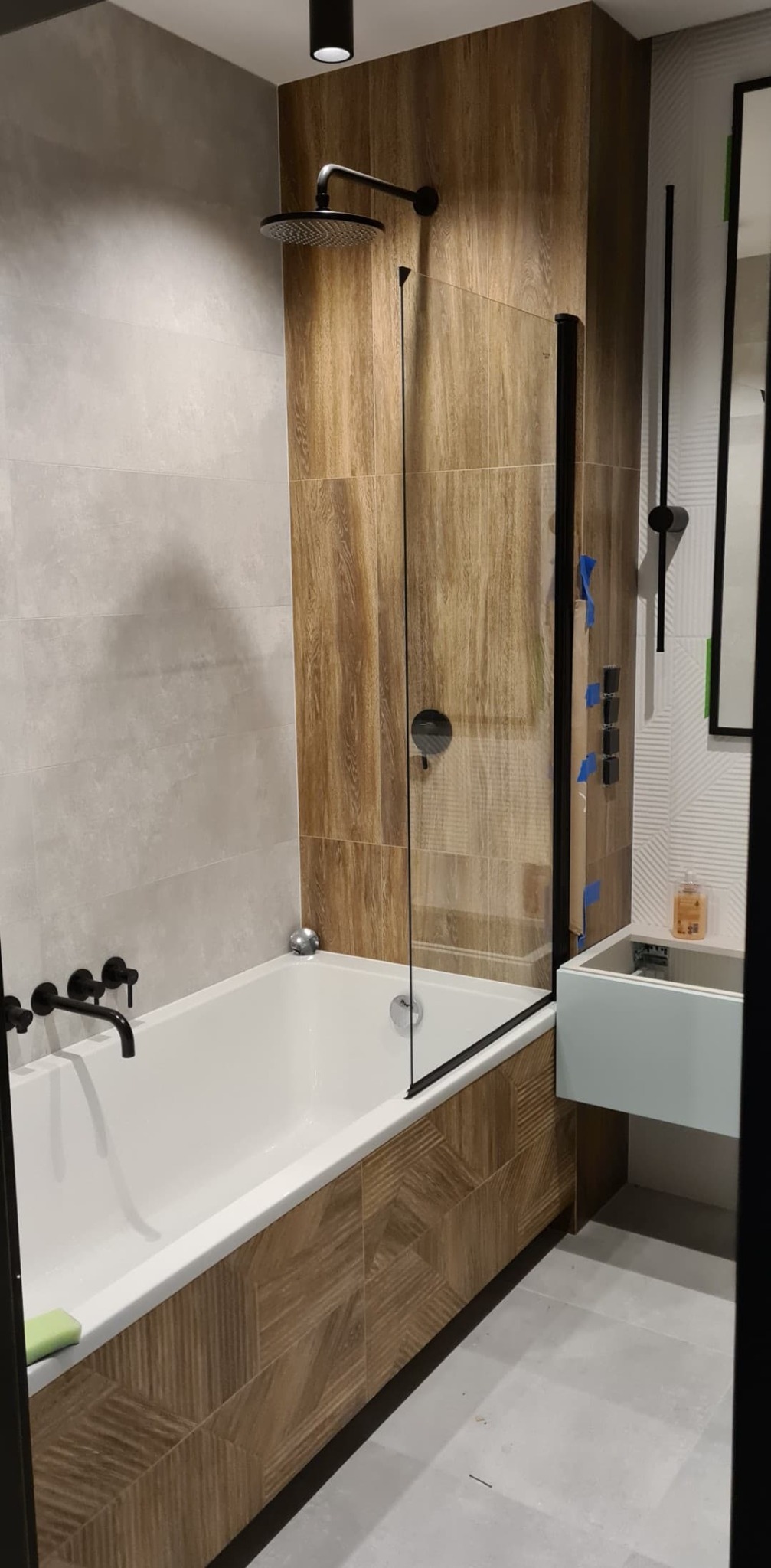 Nowoczesna łazienka po remoncie: wanna z parawanem, drewnopodobne płytki, armatura w czarnym macie i minimalistyczna umywalka.