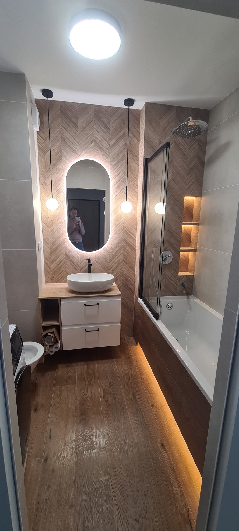 Nowoczesna łazienka po remoncie: drewnopodobna podłoga, ściany z płytek w jodełkę, owalne lustro z podświetleniem LED i wanna z czarną szybą.