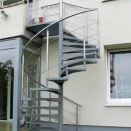 Zewnętrzne, spiralne schody z metalu, prowadzące na balkon, z widoczną konstrukcją i poręczami ze stali nierdzewnej, umieszczone przy budynku z oknami.