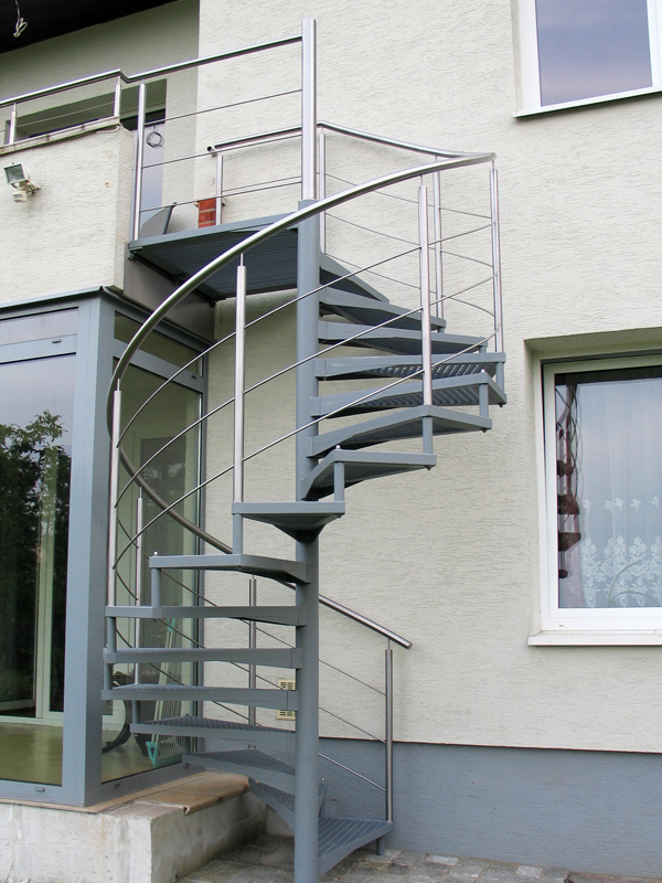 Zewnętrzne, spiralne schody z metalu, prowadzące na balkon, z widoczną konstrukcją i poręczami ze stali nierdzewnej, umieszczone przy budynku z oknami.