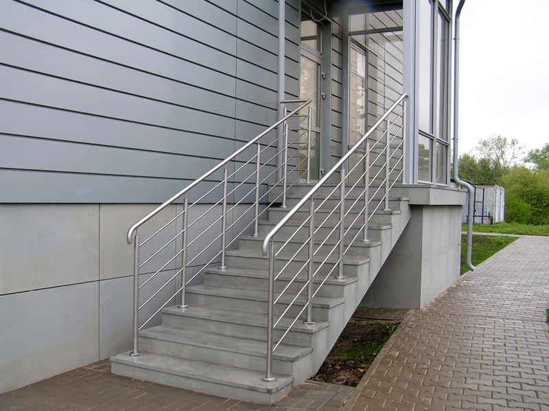 Zewnętrzne schody betonowe z metalową balustradą o minimalistycznym designie, prowadzące do budynku z szarą elewacją panelową i przeszklonym wejściem.