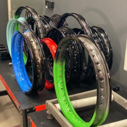 MDA DAMIAN MALISZEWSKI - Rzędy kolorowych, metalowych obręczy z otworami, prawdopodobnie felgi rowerowe, po procesie malowania, ustawione na stole warsztatowym w pracowni.