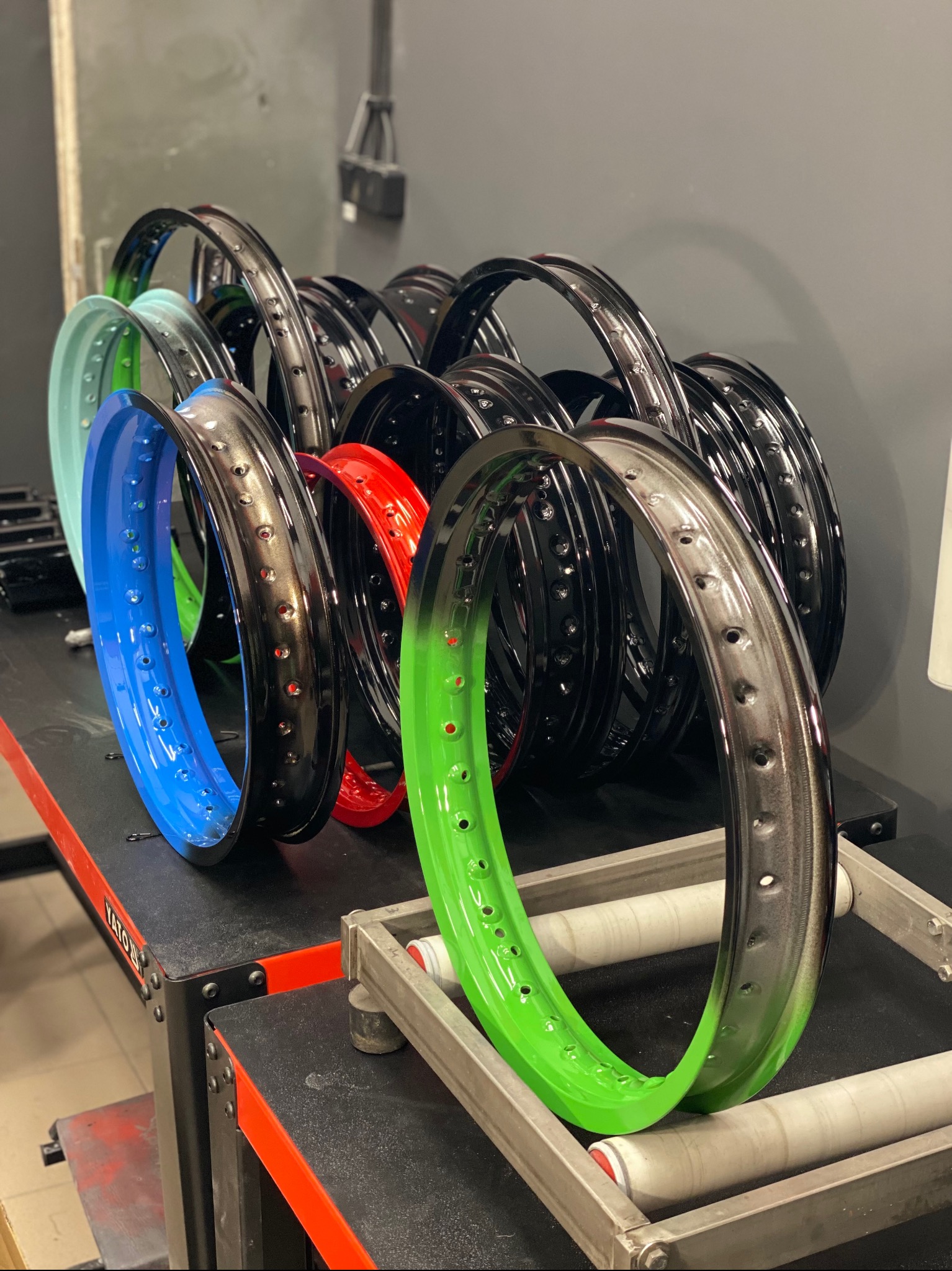 Rzędy kolorowych, metalowych obręczy z otworami, prawdopodobnie felgi rowerowe, po procesie malowania, ustawione na stole warsztatowym w pracowni.