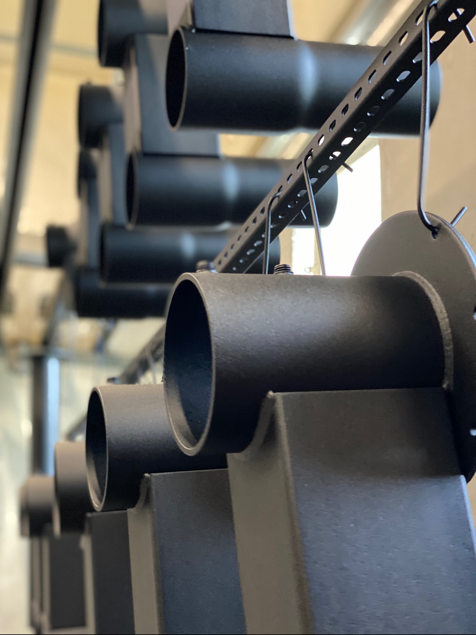 Czarne, metalowe elementy konstrukcyjne, prawdopodobnie po malowaniu proszkowym, zawieszone na metalowym stelażu, widoczne z bliska, w rzędzie.