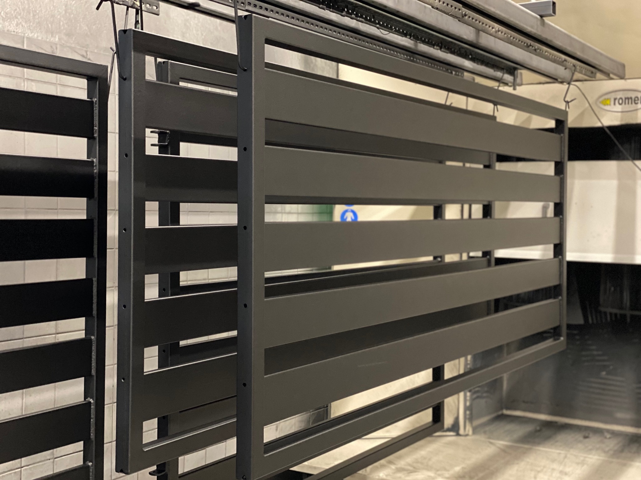 Metalowe elementy ogrodzenia, zawieszone na szynie w hali, po procesie malowania proszkowego, w kolorze ciemnoszarym, gotowe do dalszej obróbki lub transportu.