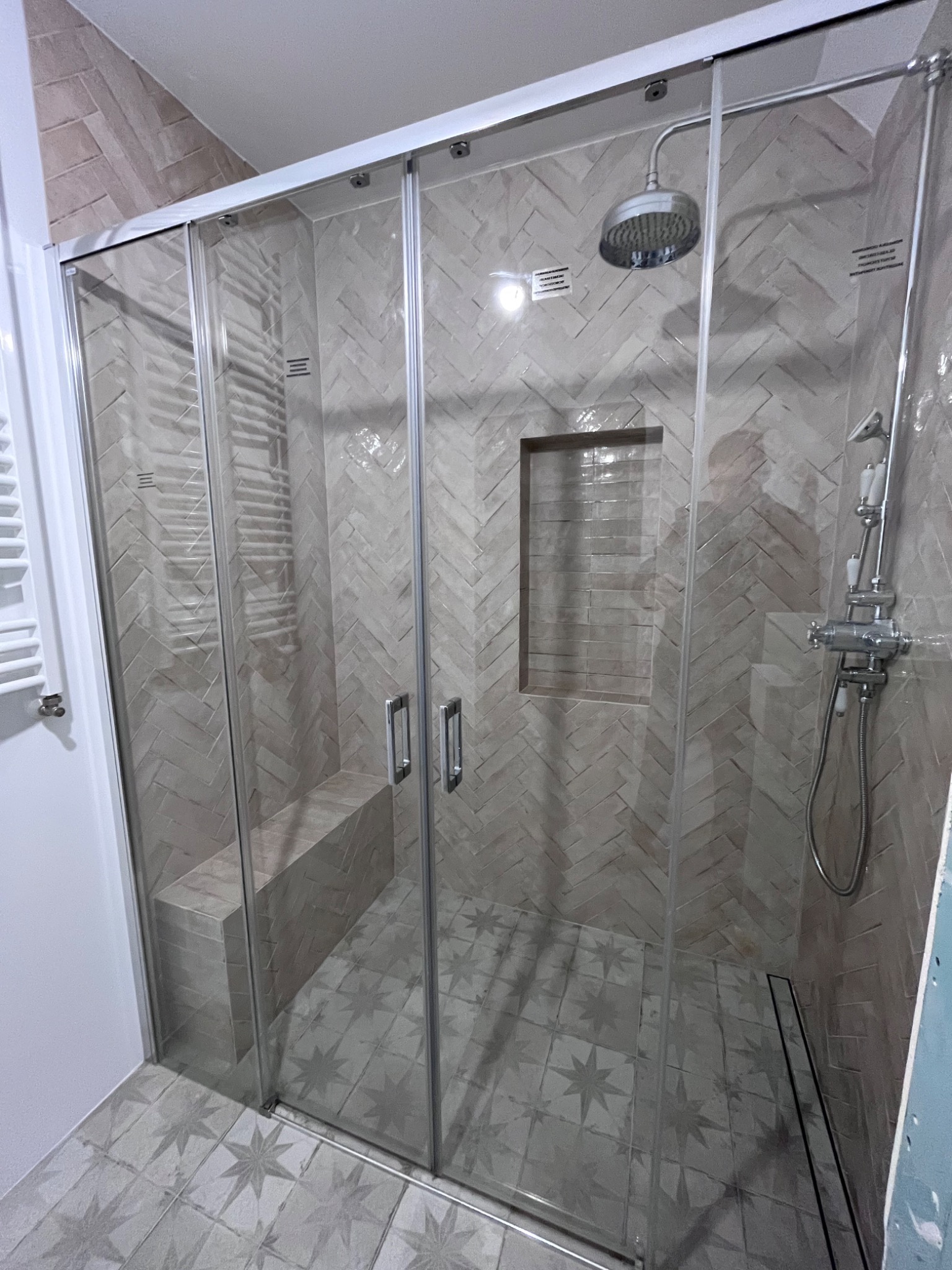 Nowoczesna kabina prysznicowa z siedziskiem, wykończona płytkami ułożonymi w jodełkę i wzorzystą podłogą. Widoczna bateria i deszczownica.