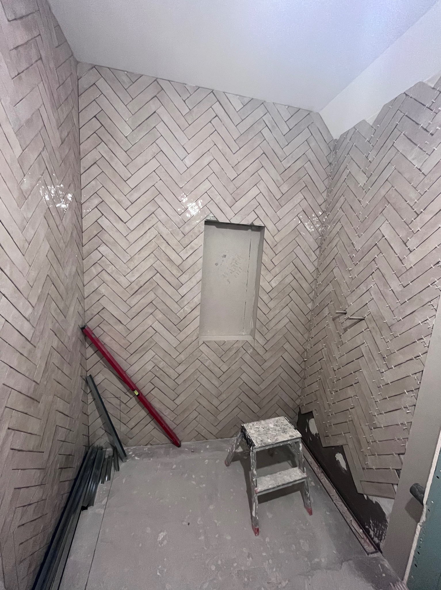 Ściany łazienki wyłożone jasnymi, błyszczącymi płytkami ułożonymi w jodełkę. Widoczny poziomica, drabinka i niedokończone fugowanie. W ścianie wnęka.