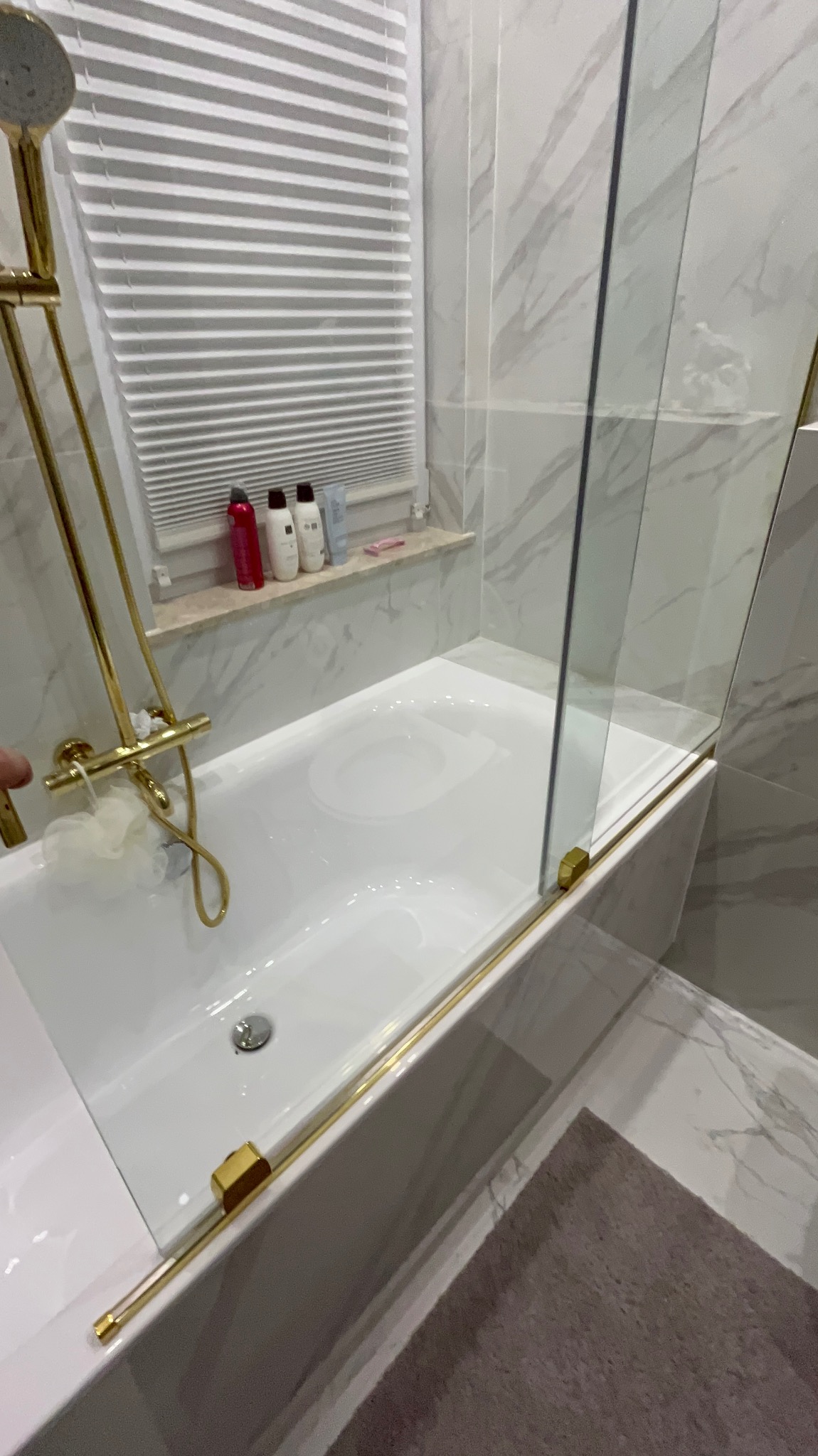 Biała wanna z parawanem nawannowym i złotą armaturą na tle marmurowych płytek. Na parapecie okiennym kosmetyki. Nowoczesna aranżacja łazienki.