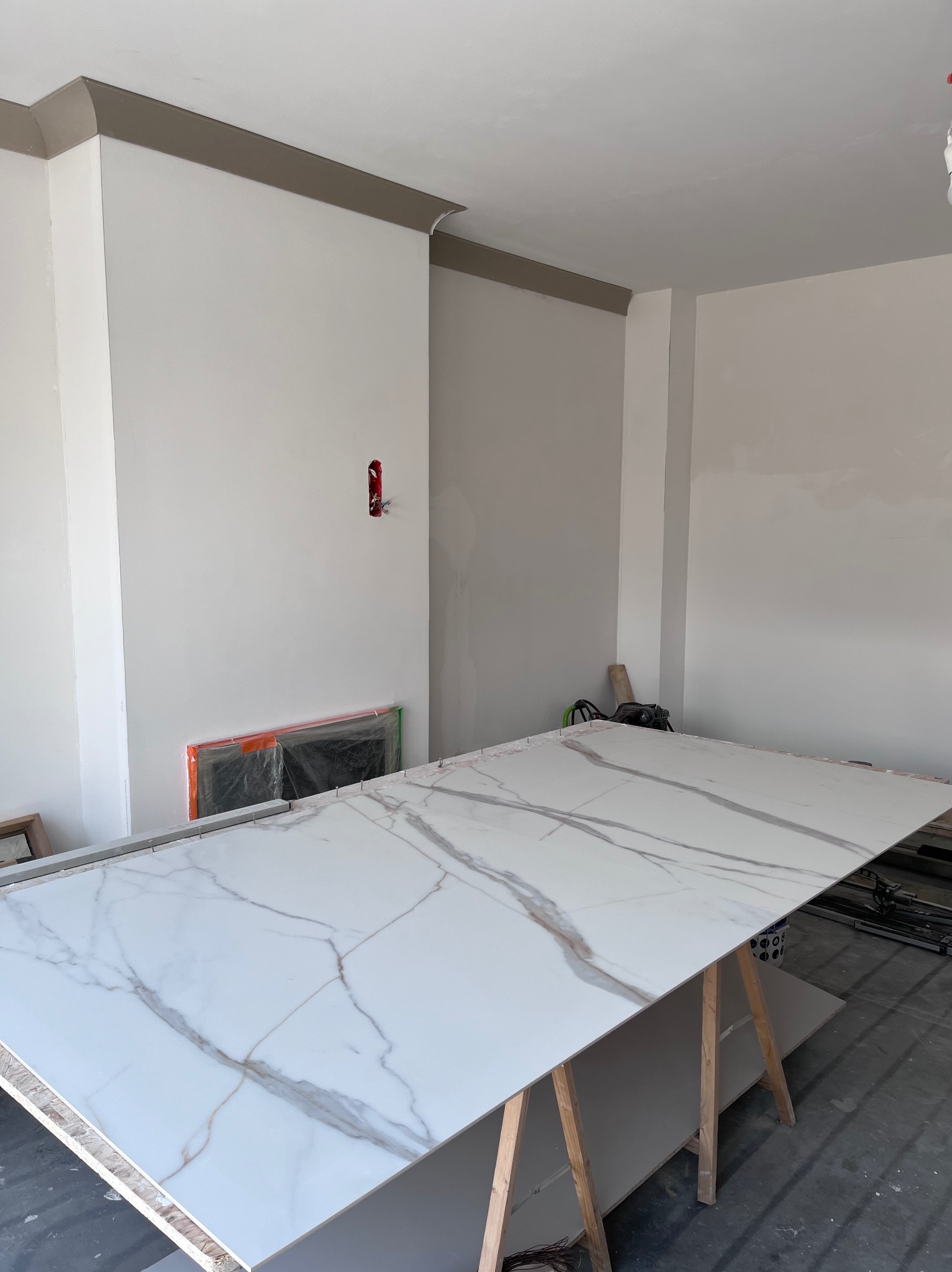 Przygotowanie płyty z marmurowym wzorem na drewnianych kozłach w jasnym wnętrzu. Widoczne narzędzia i instalacja elektryczna w ścianie.