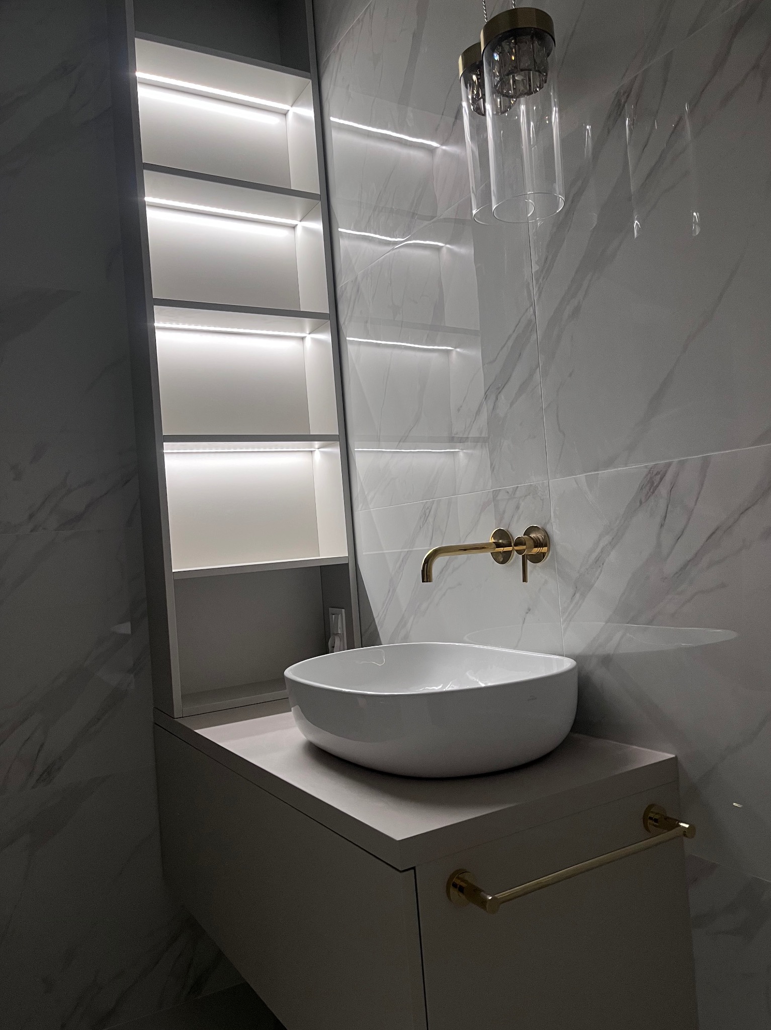 Nowoczesna łazienka z marmurowymi płytkami, okrągłą umywalką nablatową, złotą armaturą i podświetlaną szafką wnękową. Minimalistyczny design, eleganckie wykończenie.