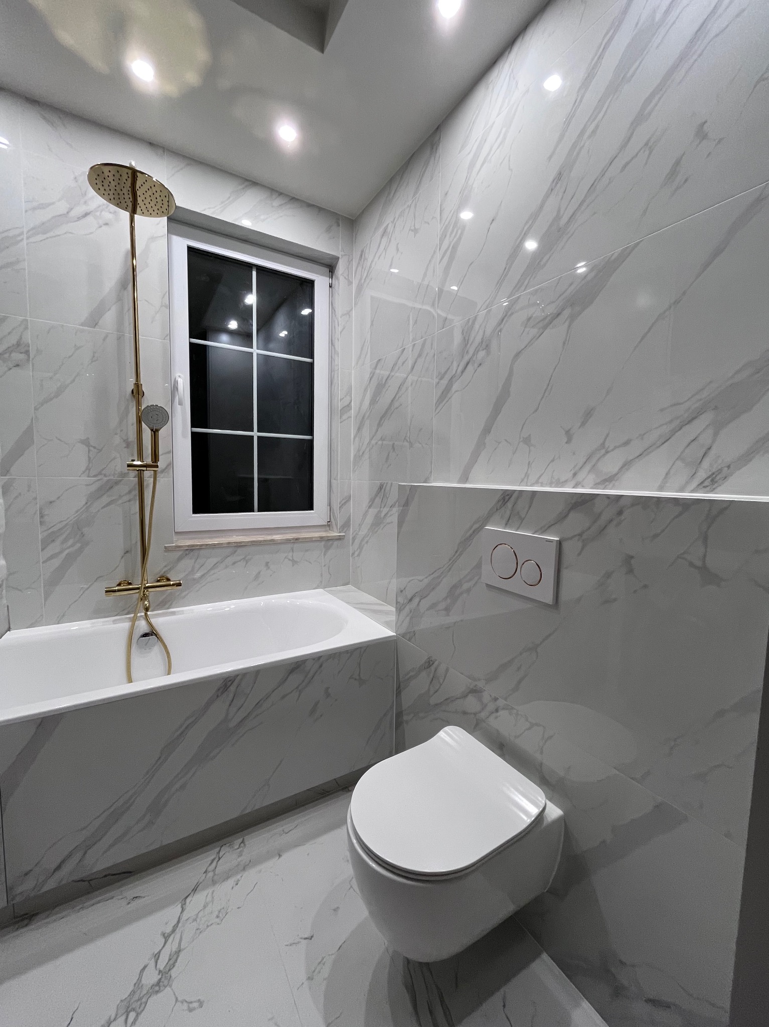 Biała łazienka z marmurowymi płytkami, złotą armaturą i podwieszaną toaletą. Nowoczesny design z oknem i punktowym oświetleniem.