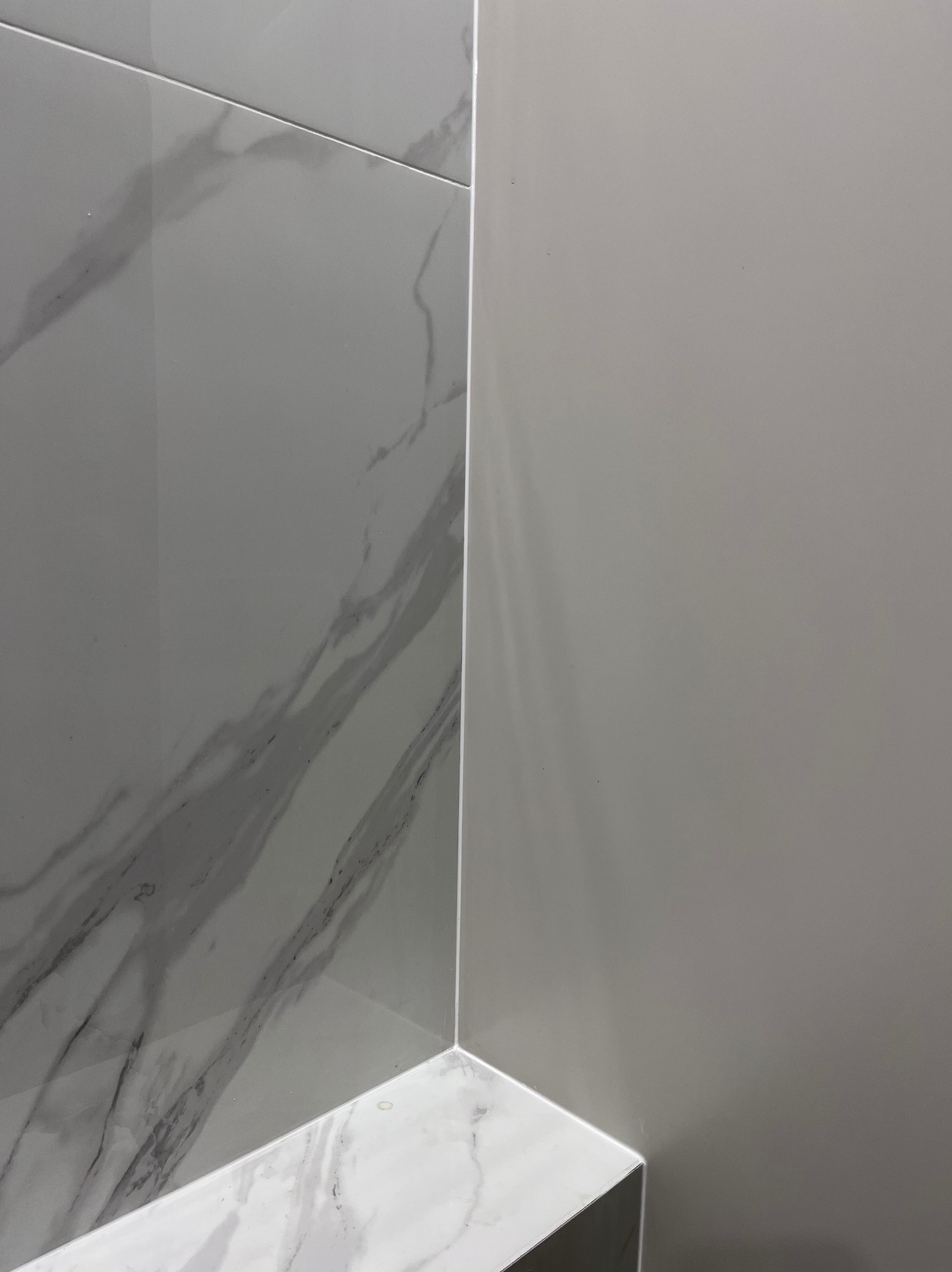 Wykończony narożnik łazienki z szarymi płytkami imitującymi marmur na ścianie i podłodze, połączonymi białą listwą. Minimalistyczny design, gładka powierzchnia.