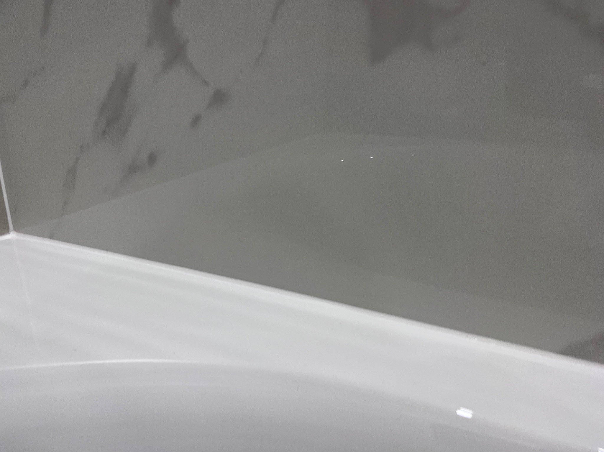 Fragment wykończonej łazienki: biała wanna z akrylu i ściana wyłożona szarymi płytkami imitującymi marmur. Minimalistyczny design, gładkie powierzchnie.