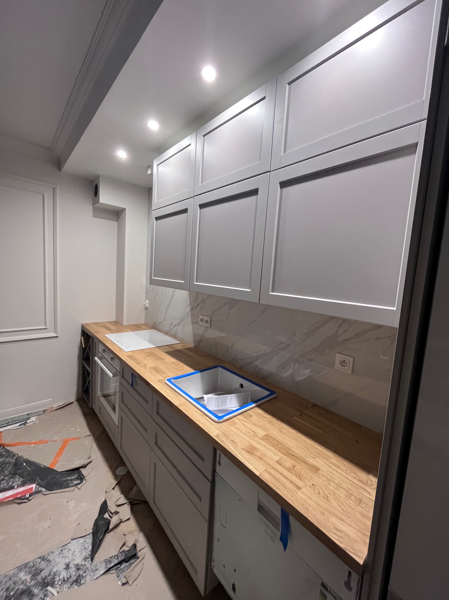 Jasna kuchnia w trakcie montażu: szare szafki z frezowanymi frontami, drewniany blat, marmurowe płytki na ścianie. Widoczne zabezpieczenia podłogi.