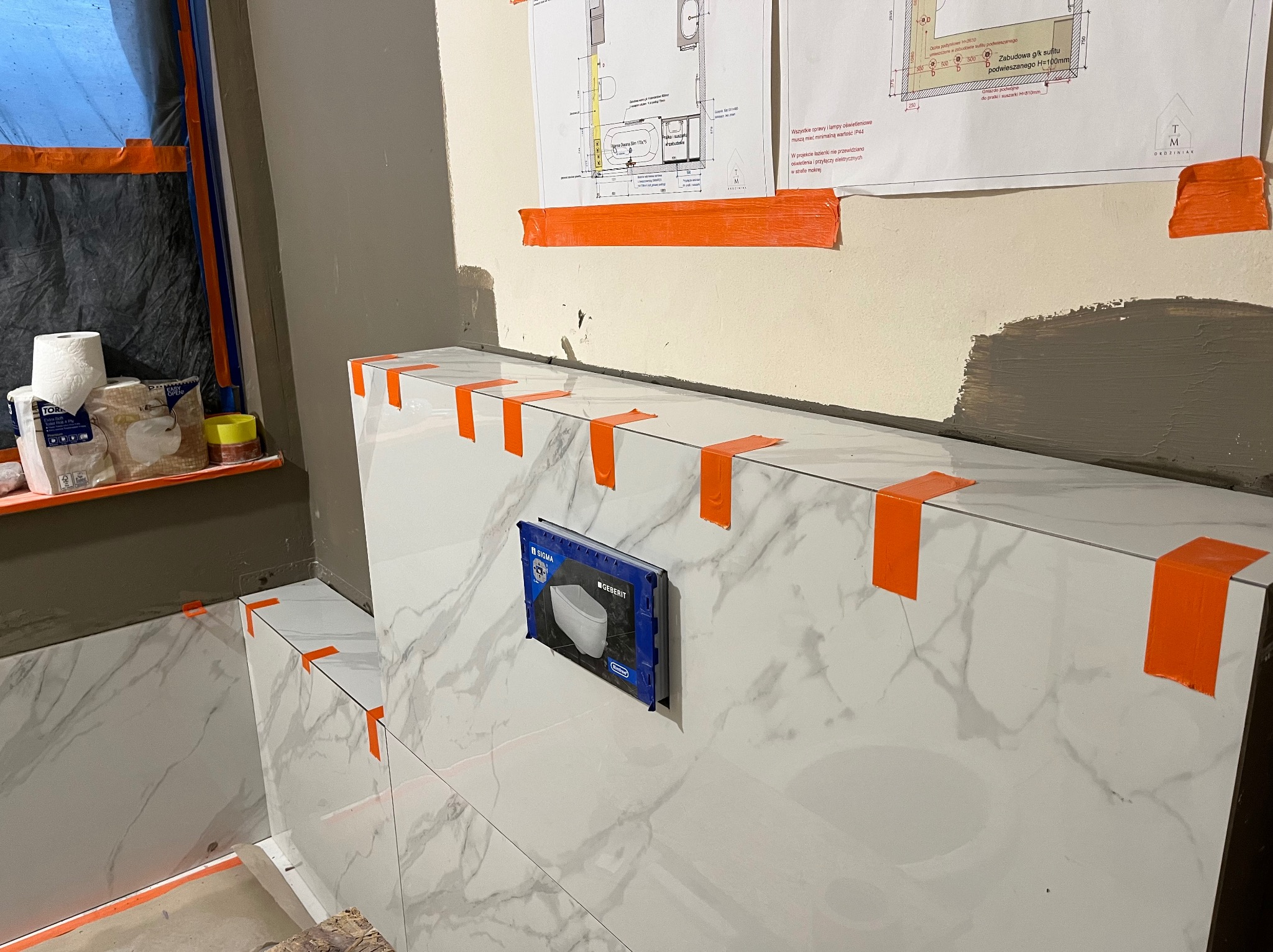 Montaż białych płytek imitujących marmur w łazience, zabezpieczonych pomarańczową taśmą. Widoczna instalacja podtynkowa Geberit i plany łazienki na ścianie.
