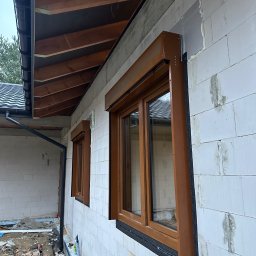 OknoMax Maciej Szewczuk - Montaż brązowych rolet zewnętrznych na oknach w Chełmie. Widoczny fragment elewacji budynku z białego bloczka i elementy orynnowania.