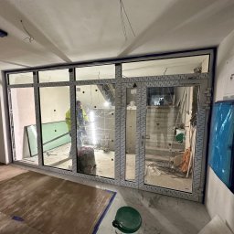 OknoMax Maciej Szewczuk - Montaż dużych okien z ramą PCV w surowym wnętrzu. Widoczne narzędzia i elementy wykończeniowe. Praca w toku, z dbałością o detale.