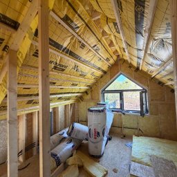 OknoMax Maciej Szewczuk - Dach domu szkieletowego w trakcie izolacji: widoczne drewniane elementy konstrukcyjne, folia paroizolacyjna Eurovent, okno trójkątne i rolki wełny mineralnej URSA.