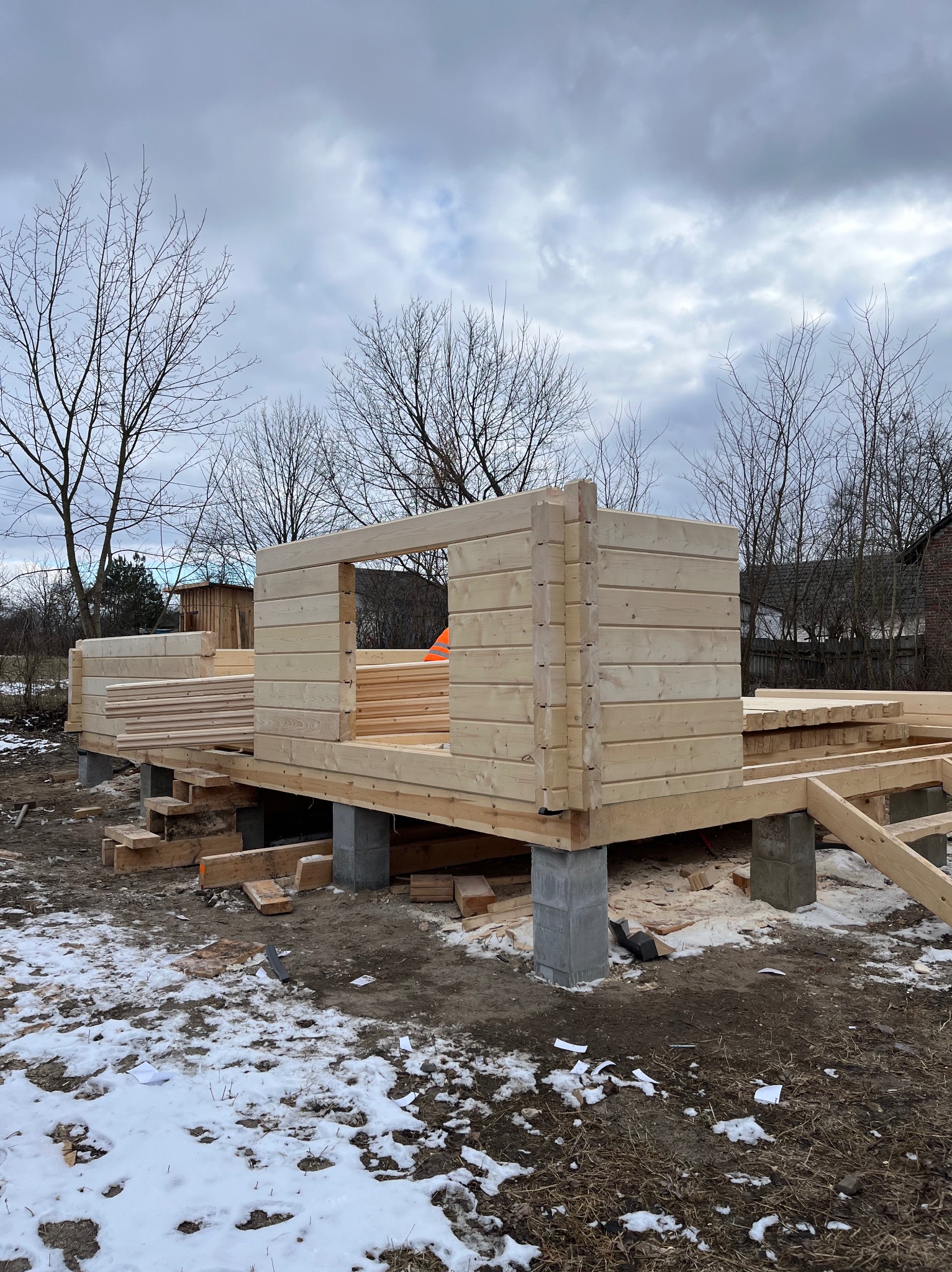 Surowy stan domu szkieletowego w trakcie budowy. Widoczne drewniane ściany i podłoga na betonowych fundamentach, zima w tle. Częściowo ułożone deski.