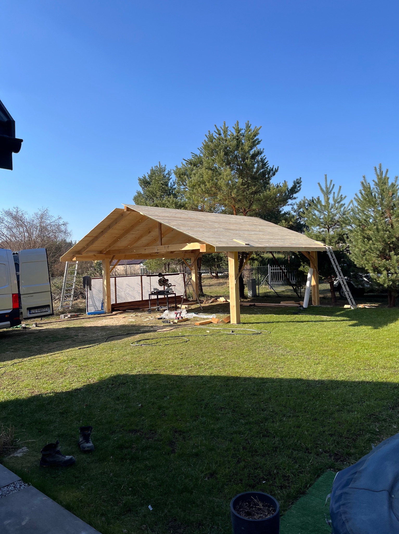 Drewniana konstrukcja zadaszenia w trakcie budowy na trawniku, z narzędziami i drabiną. Błękitne niebo w tle. Widoczne buty robocze i samochód dostawczy.