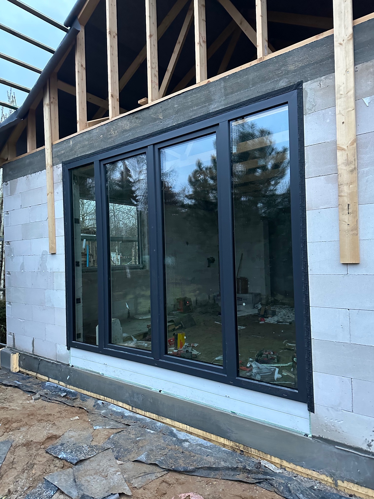 Ciemne, panoramiczne okno w surowym stanie domu z białego bloczka. Widoczne drewniane elementy konstrukcji dachu i odbicie drzew w szybach. Etap budowy.