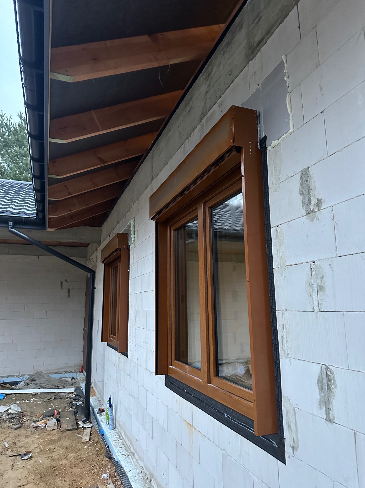 Montaż brązowych rolet zewnętrznych na oknach w Chełmie. Widoczny fragment elewacji budynku z białego bloczka i elementy orynnowania.