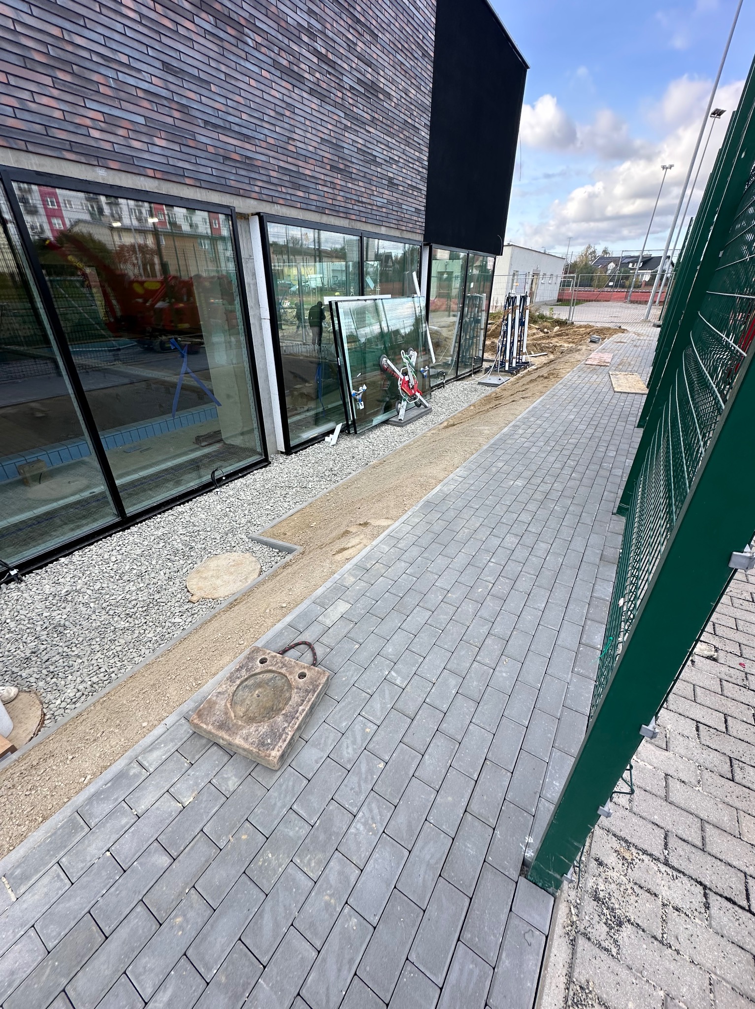 Montaż dużych okien w budynku z cegły. Widoczne okna przygotowane do instalacji, narzędzia i fragment chodnika z kostki brukowej. Ujęcie z zewnątrz budynku.