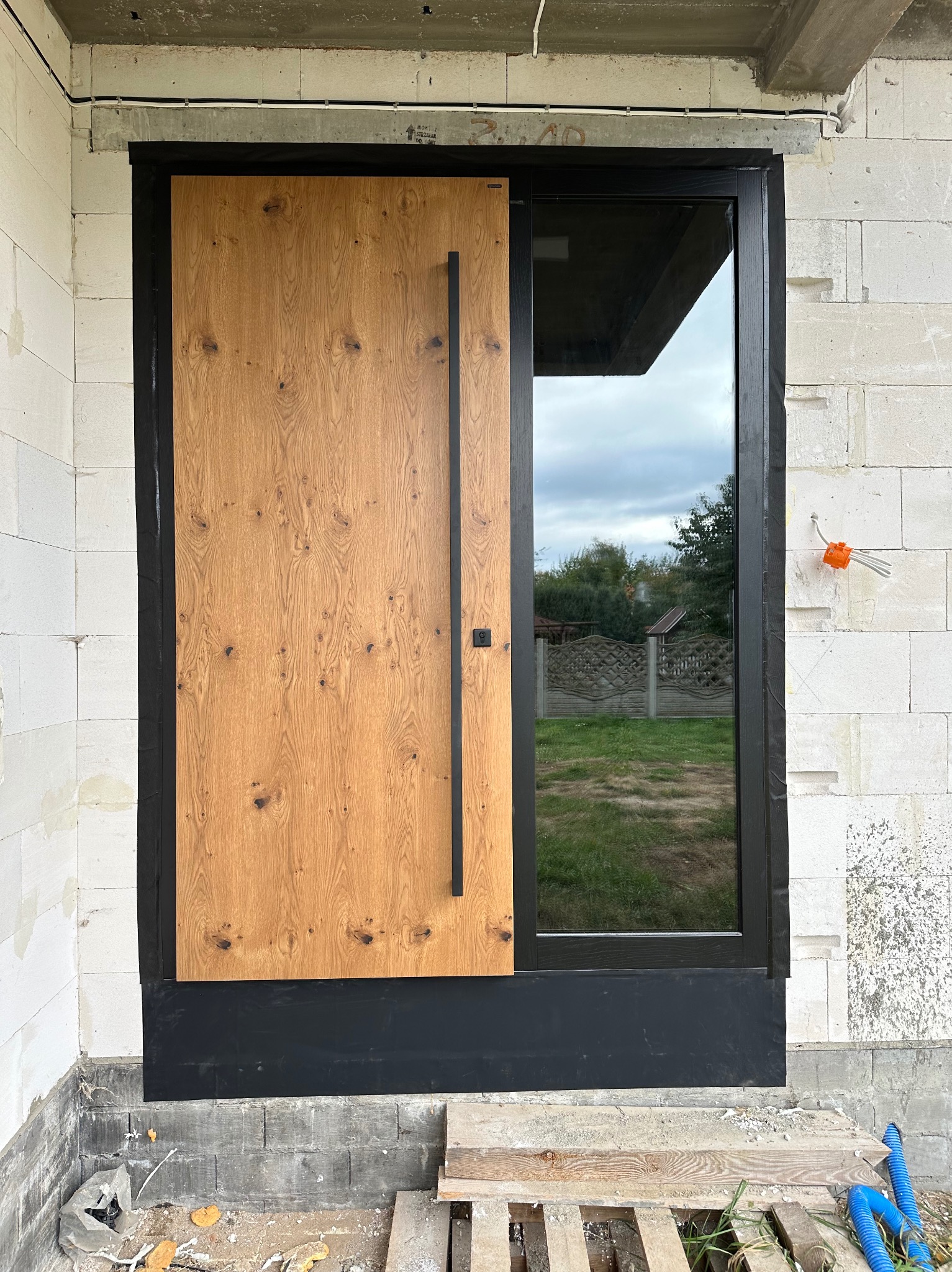Nowoczesne drzwi zewnętrzne z drewnianym panelem i czarną ramą, zamontowane w surowym murze z betonu komórkowego. Widoczne elementy instalacji elektrycznej.