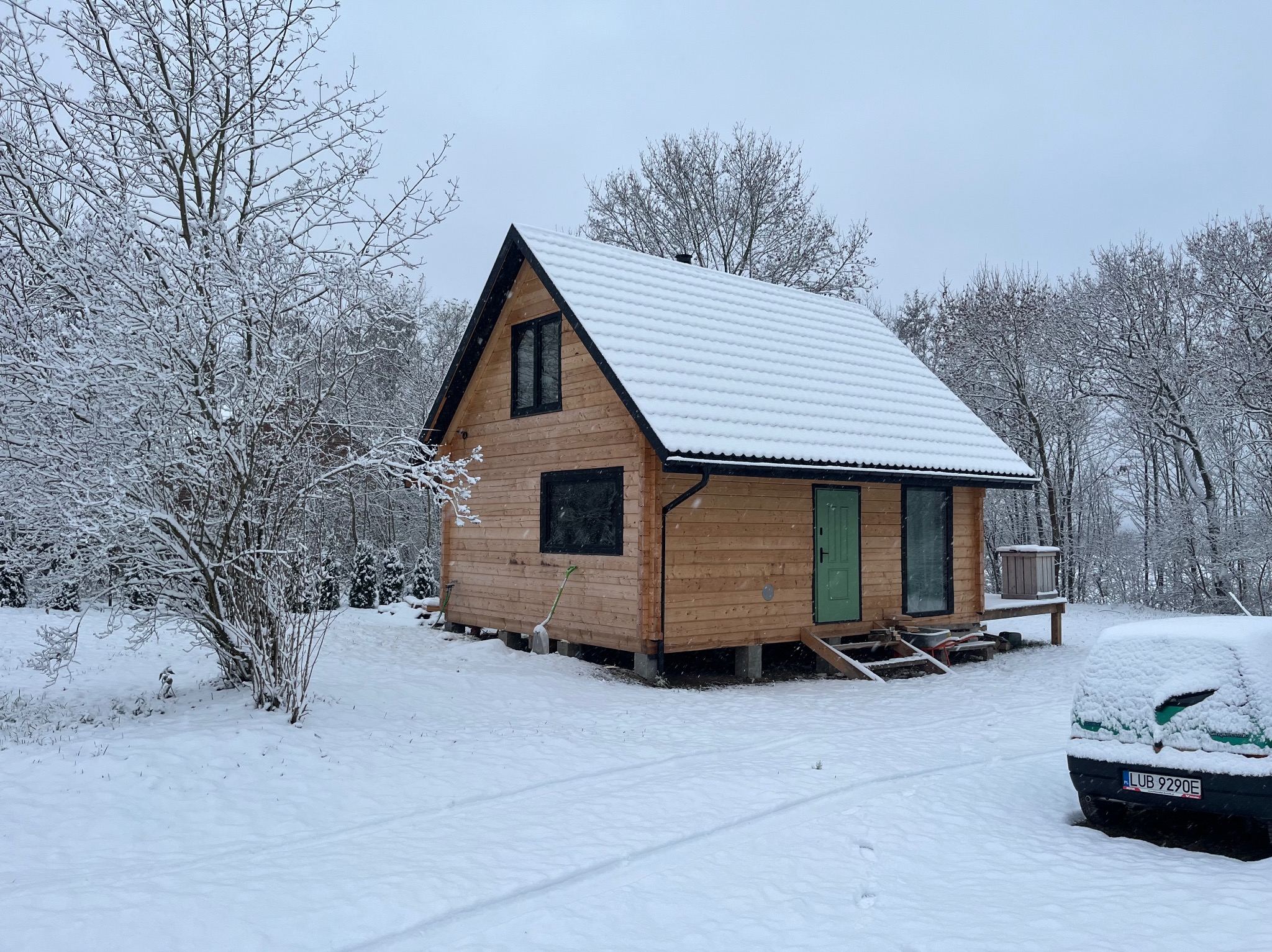 Zimowy krajobraz z drewnianym domem szkieletowym na podwyższeniu, pokrytym śniegiem. Zielone drzwi wejściowe i zasypany śniegiem samochód w tle. Śnieżne drzewa.