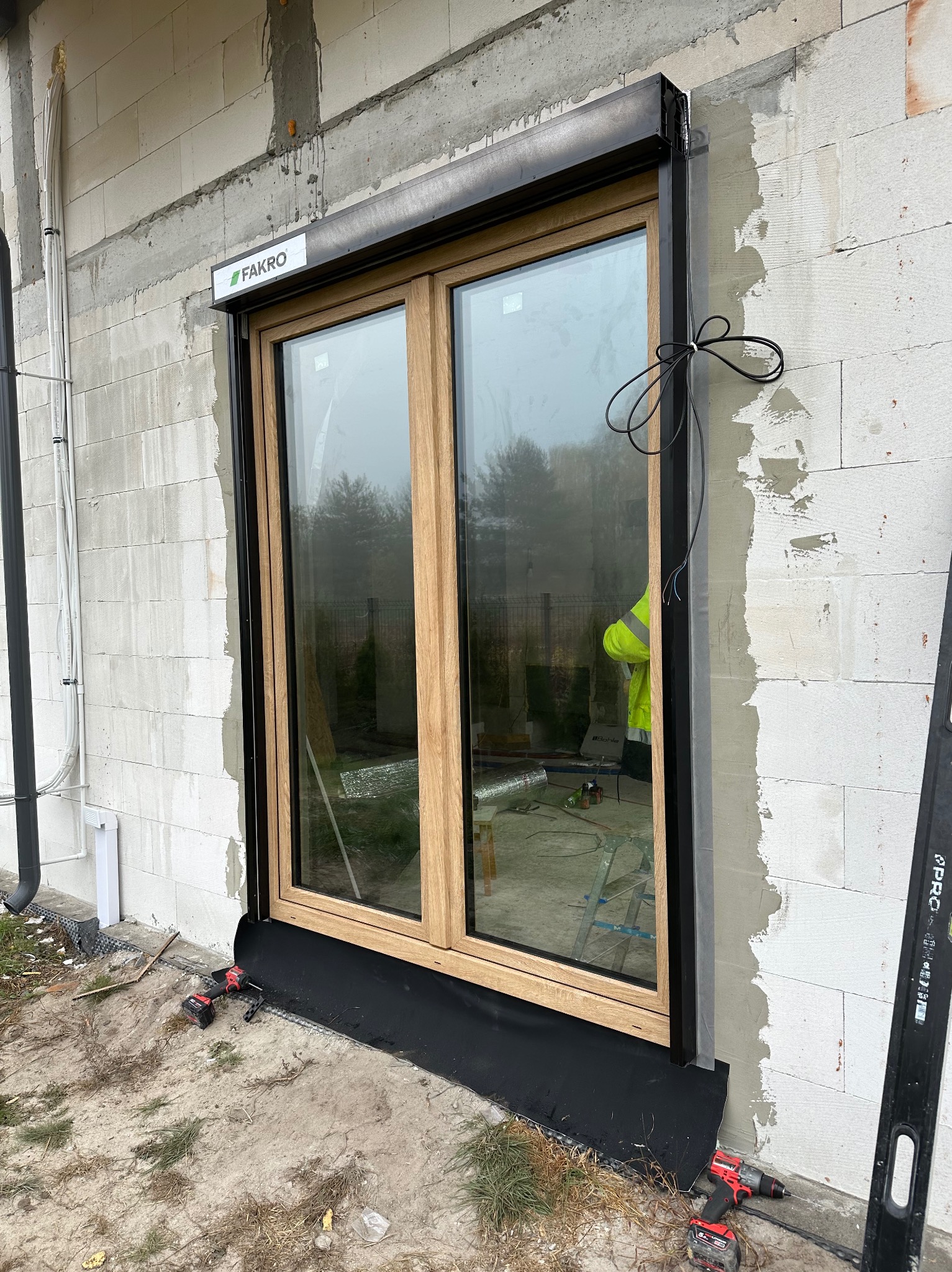 Montaż okna z roletą zewnętrzną marki Fakro w surowym murze z betonu komórkowego. Widoczne narzędzia i przewody. Okno z drewnianą ramą i czarną roletą.