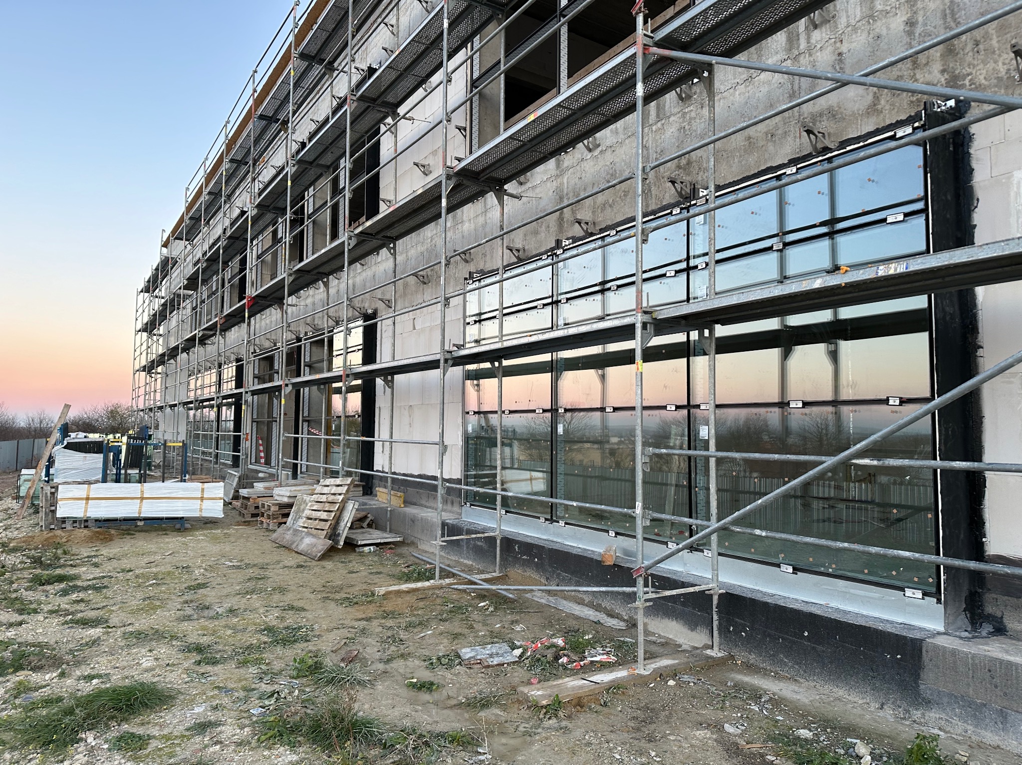 Montaż okien na surowej elewacji budynku w trakcie budowy, widoczne rusztowania i przygotowane pakiety okienne. Okna z czarnymi ramami odbijają zachodzące słońce.