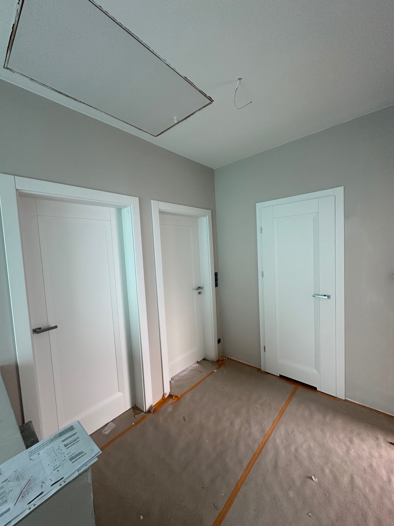 Trzy białe, nowoczesne drzwi wewnętrzne z czarnymi i srebrnymi klamkami, zamontowane w szarym pomieszczeniu. Na podłodze folia ochronna i dokumentacja.