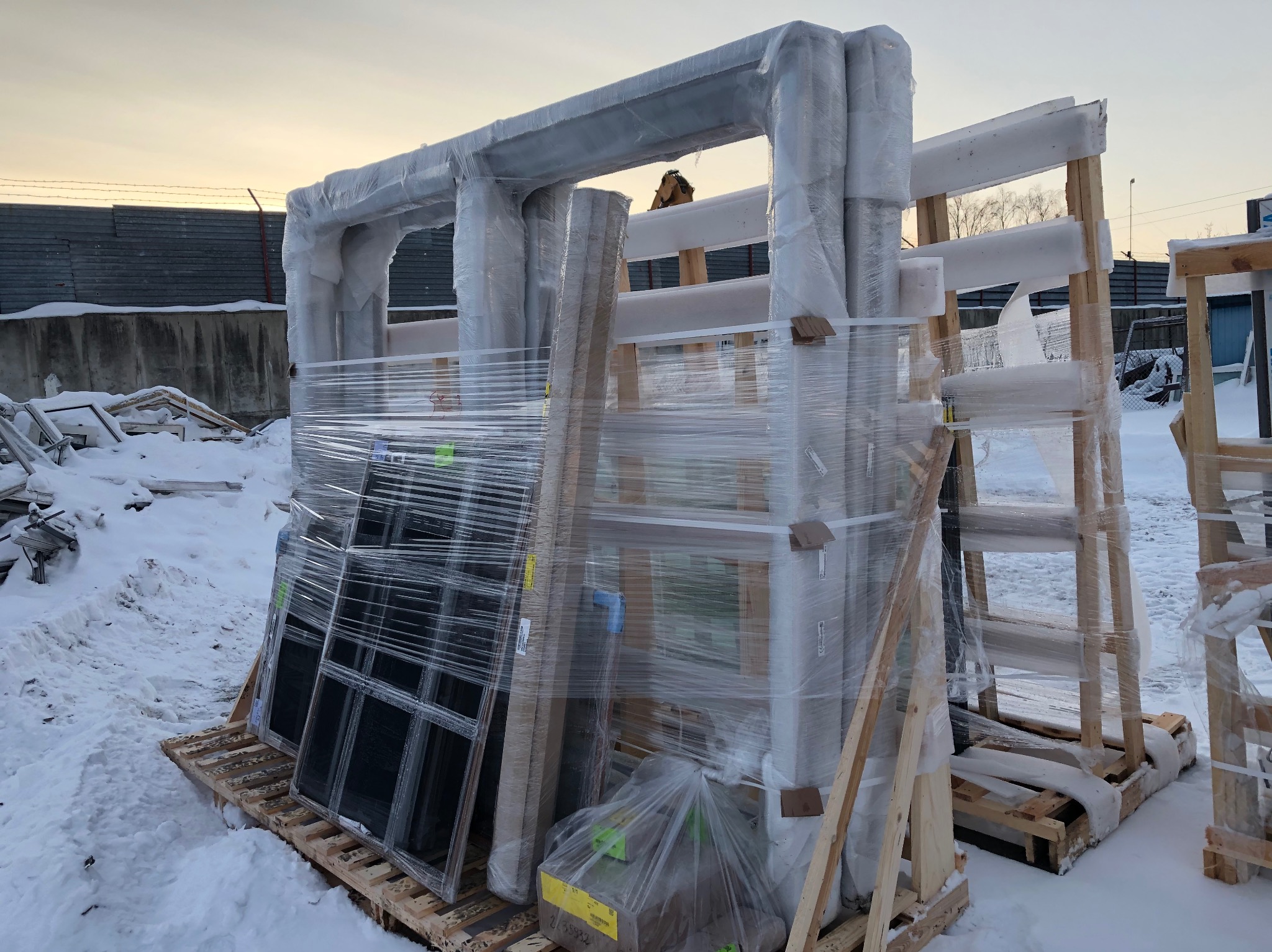 Zabezpieczone folią okna i ramy okienne, ułożone na paletach w zimowej scenerii. Widoczne drewniane podpory i śnieg. Przygotowane do transportu lub montażu.